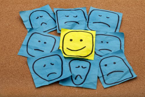 positive attitude or optimism concept - happy smiley face on yellow sticky note surrounded by sad unhappy blue faces