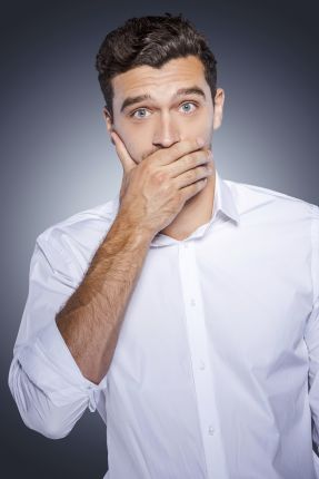 I will keep a secret! Surprised young man in white shirt covering mouth with hand and looking at camera while standing against grey background