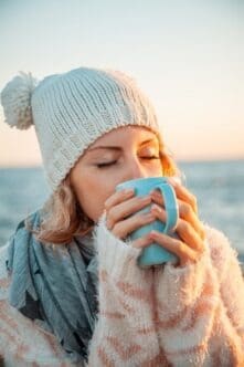 Cute young european woman in knitted clothes drinks coffee on the beach at sunset